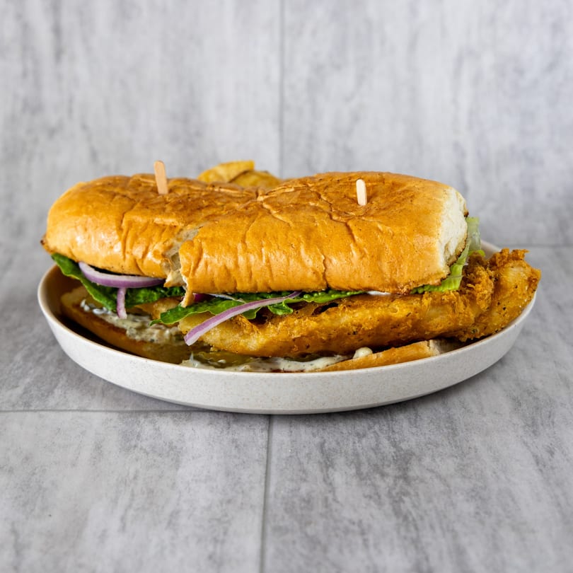 A large fried fish po'boy sandwich with lettuce, red onion, and sauce on a white plate.