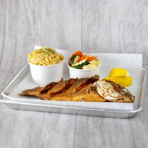 Fried whole fish with pasta, mixed vegetables, and lemon wedges on a metal tray.