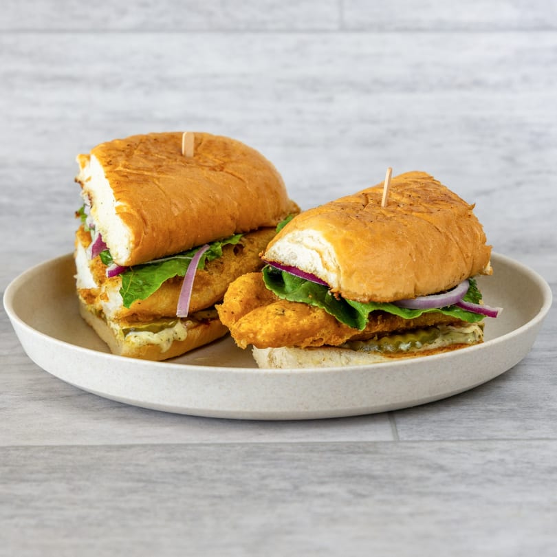 Two Uncle Mac's Kitchen fried fish sandwiches with lettuce, red onion, and pickles on a plate.